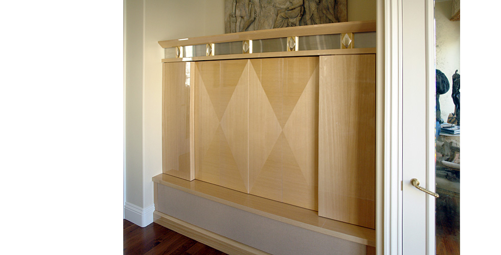 Entertainment center features sterling silver, gold and brushed stainless steel ornamental details across the top. The sliding television doors are accentuated with a harlequin patterned grain, accented again in glossy taywood cabinetry. 
